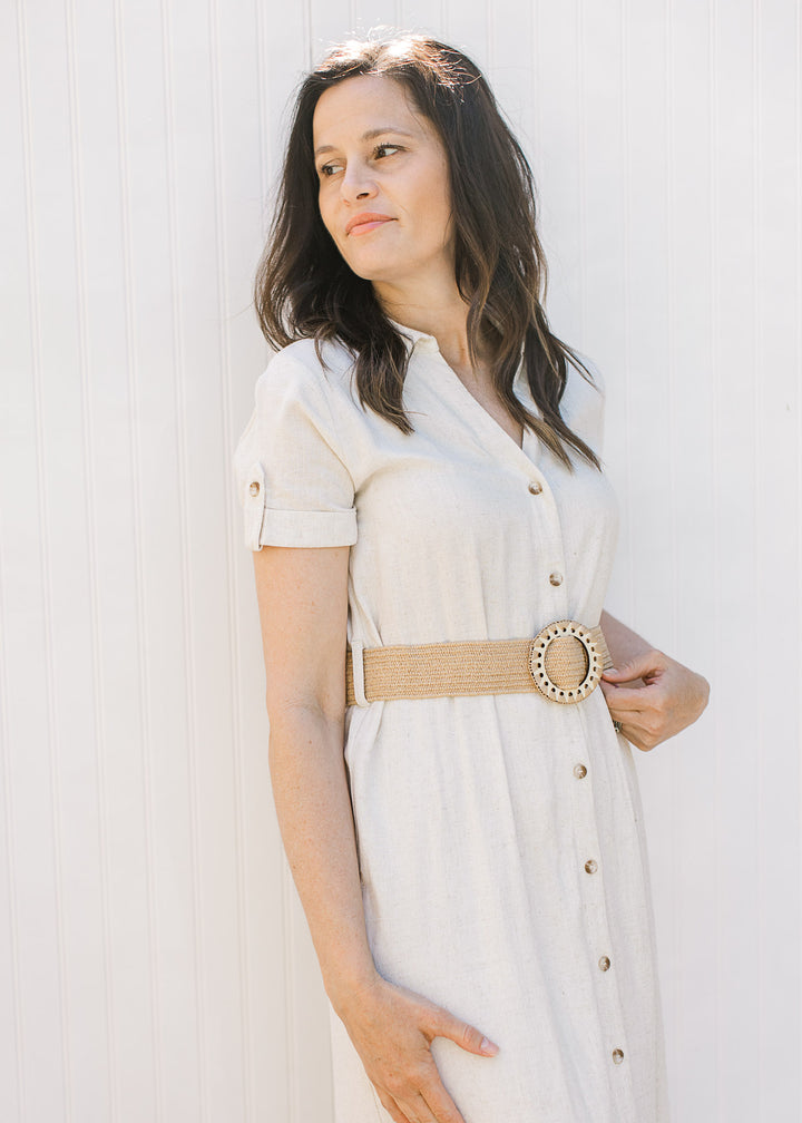 Model wearing a sandy linen dress with button front, collar, short sleeves with clasp and belt.