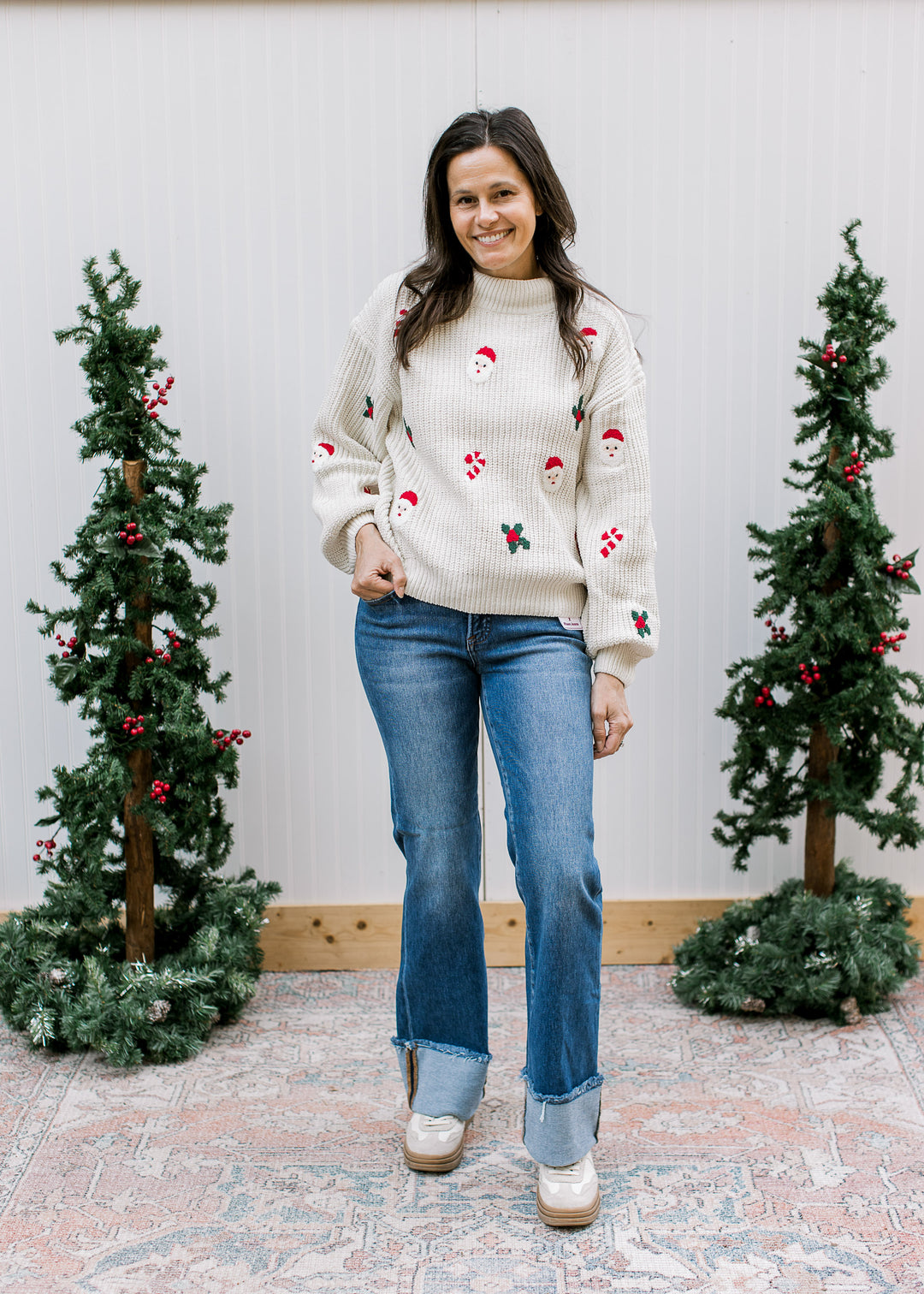 Full-length outfit with cream Santa Christmas sweater, cuffed wide leg jeans and cream shoes, beside Christmas trees.