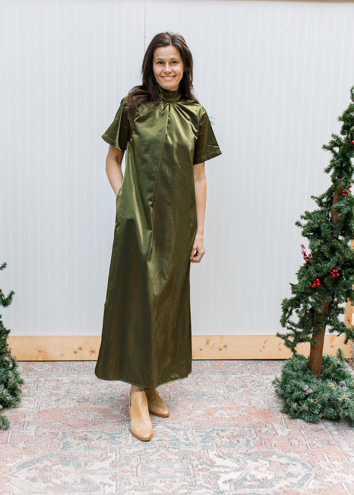 Full-length olive maxi dress with mock neckline and short sleeves, styled with tan ankle boots beside Christmas trees.