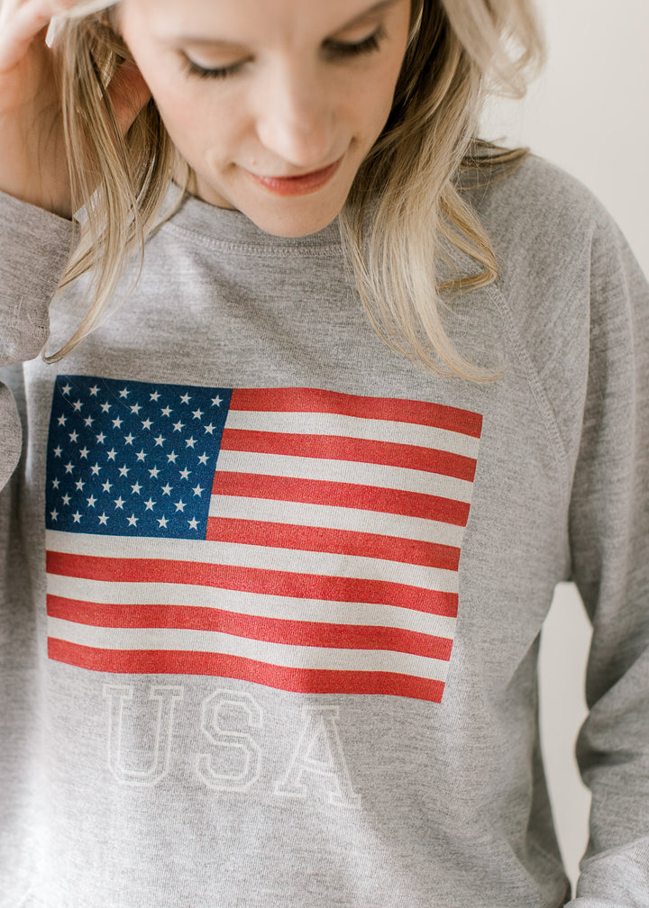 Close up of imprinted USA flag and lettering on a gray long sleeve top with a round neck. 
