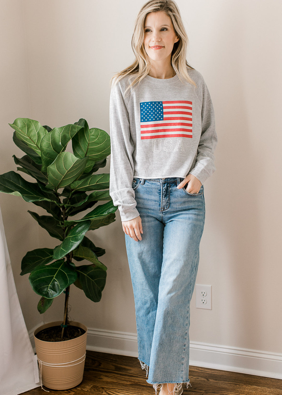 Model wearing jeans and a long sleeve slightly cropped, gray top with USA flag and lettering.