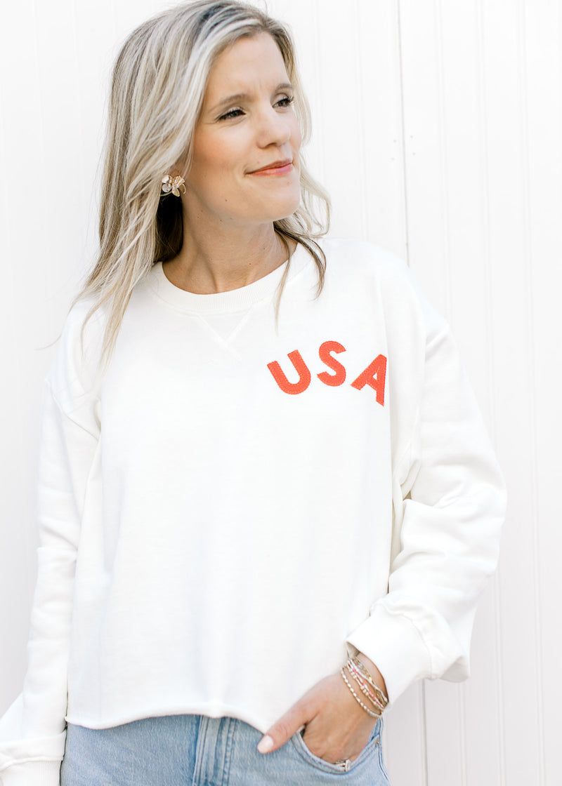 Model wearing a slightly cropped white sweatshirt with a fabric USA patch and ribbed neck. 