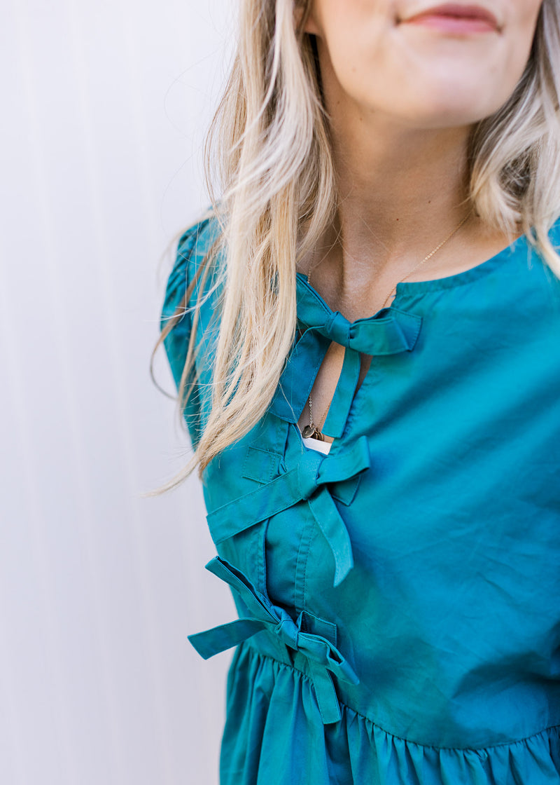 Close up of three bows down the front of a teal top with 3/4 sleeves and pleated shoulders. 