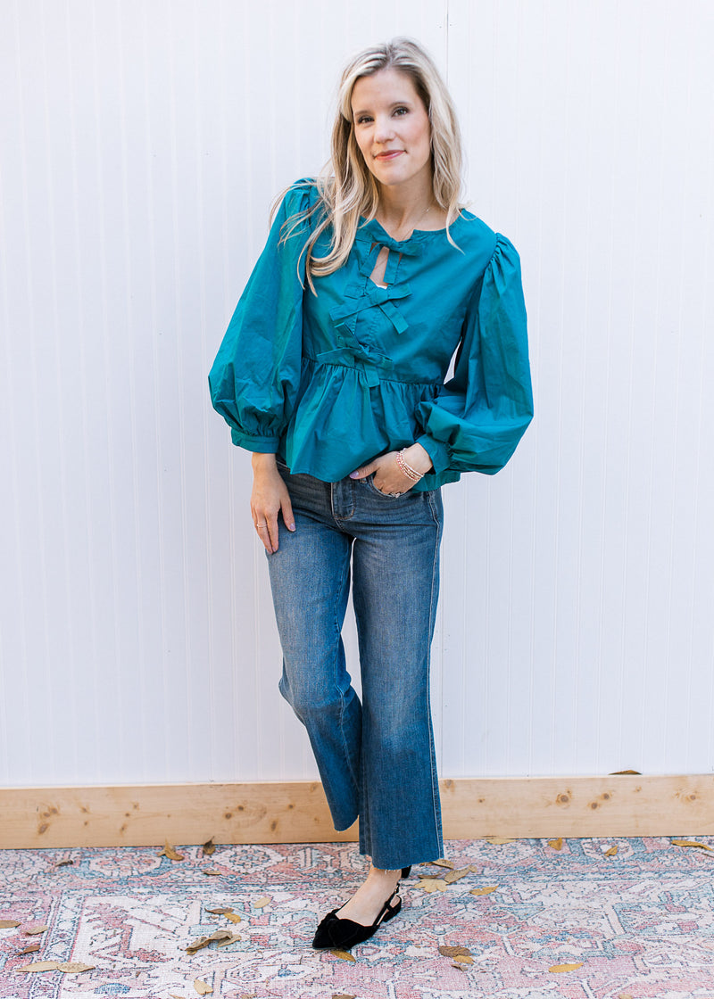 Model wearing jeans, flats and a teal top with bow details and bubble long sleeves. 