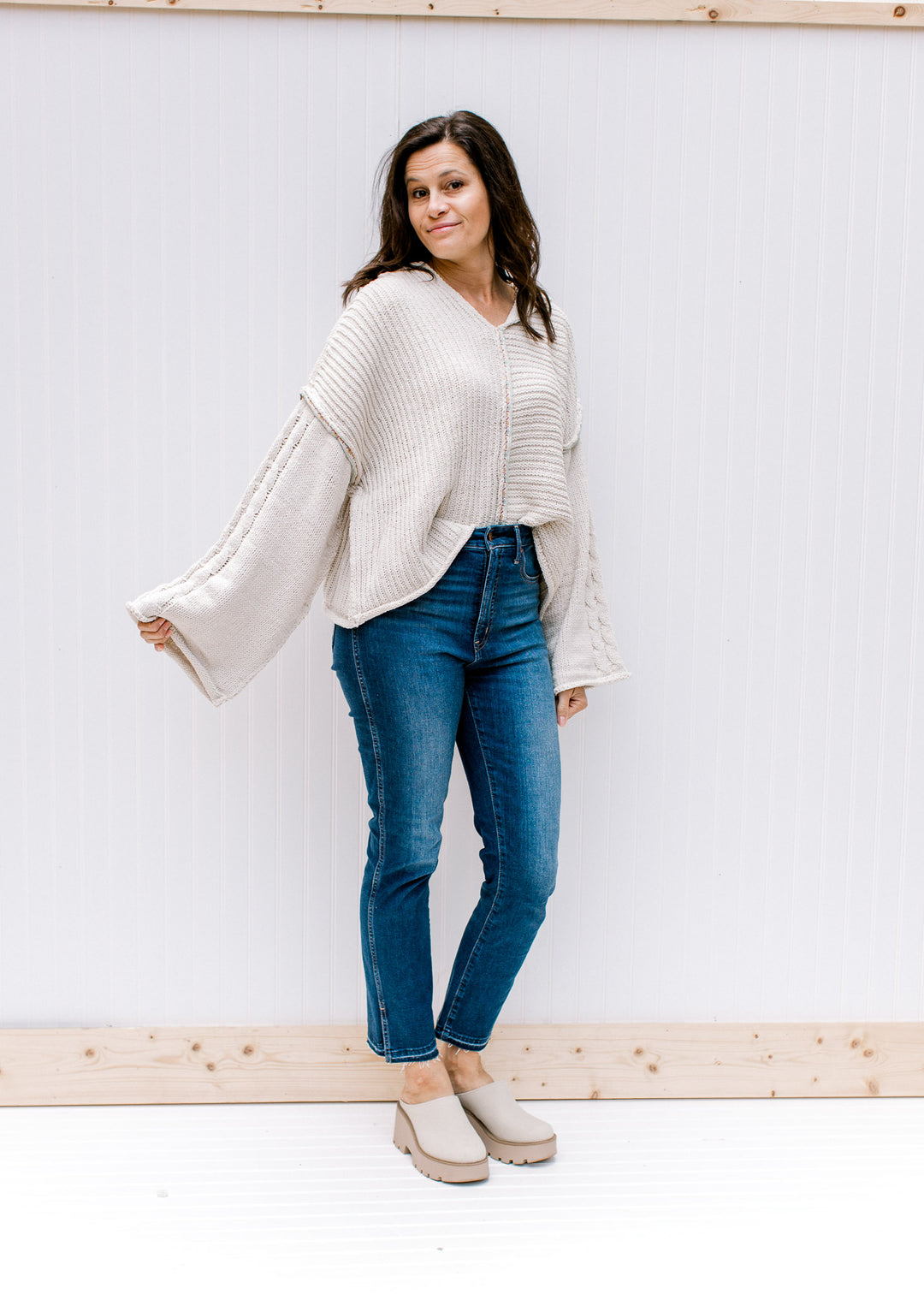 Model wearing jeans, mules and a cream cable knit sweater with kimono long sleeves and a hood. 