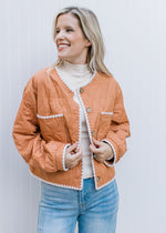 Model wearing an amber colored quilt jacket with cream embroidery and long sleeves. 