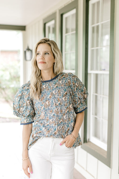 Model wearing a tan top with blue and brown floral, short sleeves and lace detail at cuff and neck.