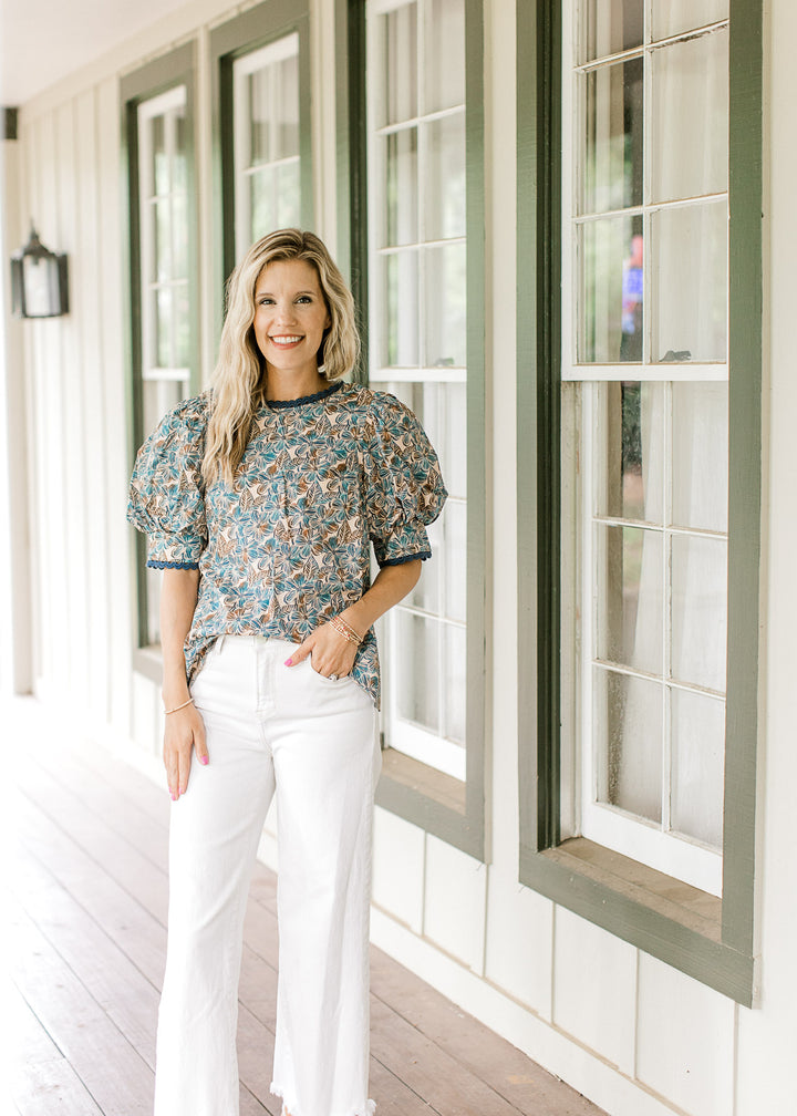 Model wearing white pants with a tan top with blue and brown floral and short puff sleeves.