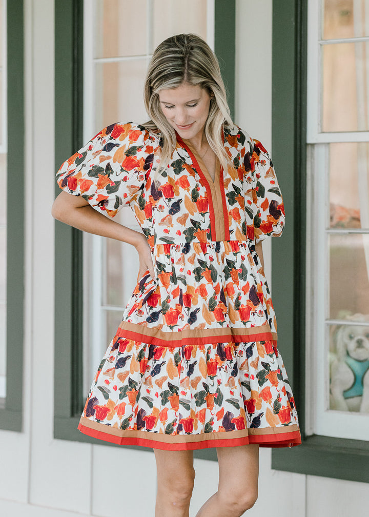 Model wearing a cream dress with fall hues, bubble short sleeves and rust and brown piping. 