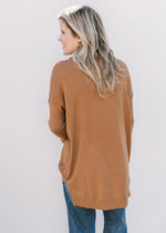 Back view of Model wearing a camel flowy sweater with long sleeves, cowl neck and split sides. 