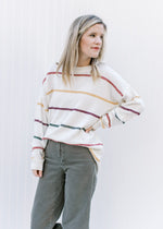 Model wearing a cream long sleeve sweater with green, burgundy, gold and red stripes.