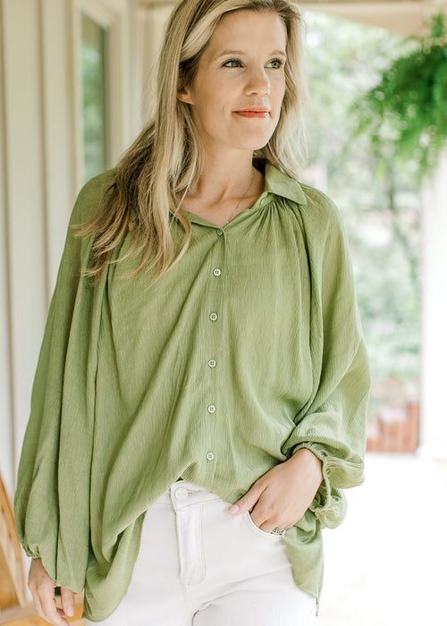 Model wearing an avocado green top with button front, collar and long sleeve with elastic band.