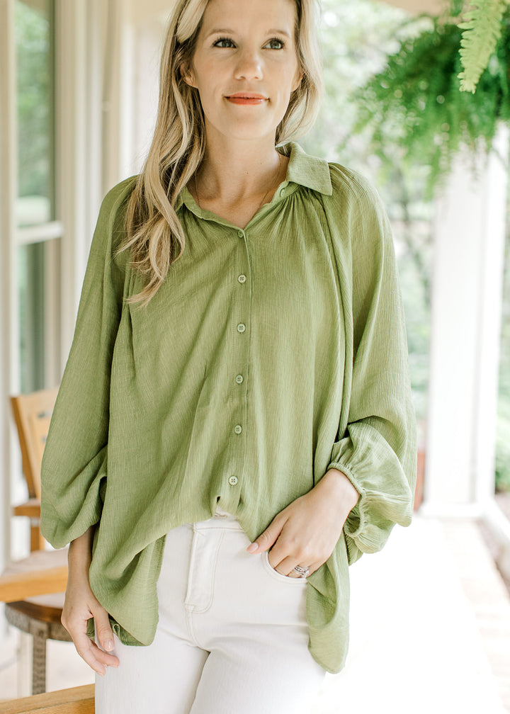 Model wearing an avocado green top with button front and pleating at shoulder of long sleeve.