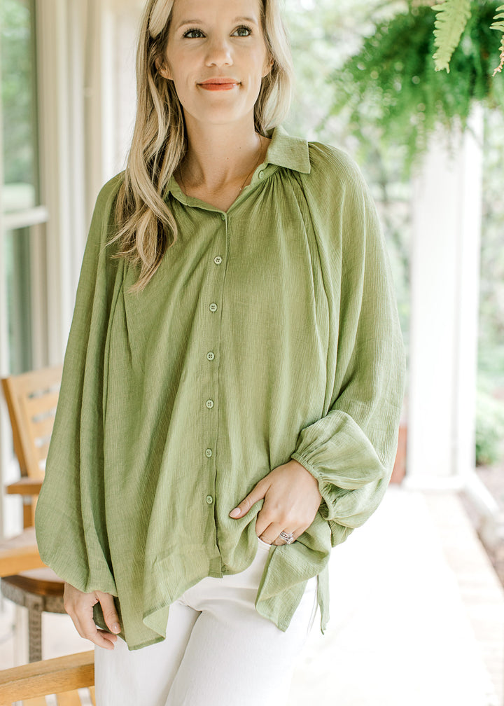 Model wearing an avocado green top with pleated shoulders and long sleeve with elastic band.