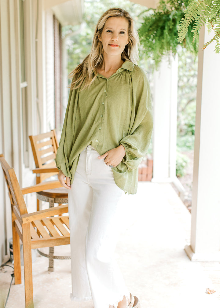 Model wearing white pants, sandals and an avocado green top with button front and long sleeve.