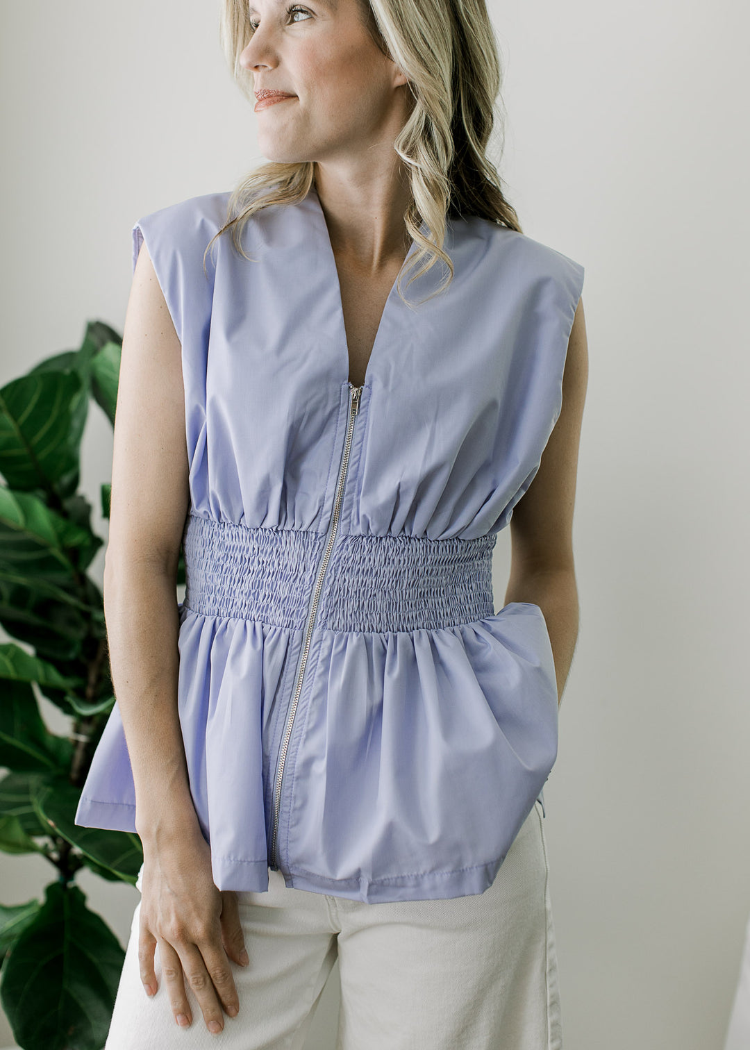 Model wearing a sleeveless baby blue top with a zipper closure, smocked waistband and v-neck. 