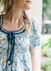 Close up of scalloped lace and faux tie on a baby blue top with cream floral and short sleeves.