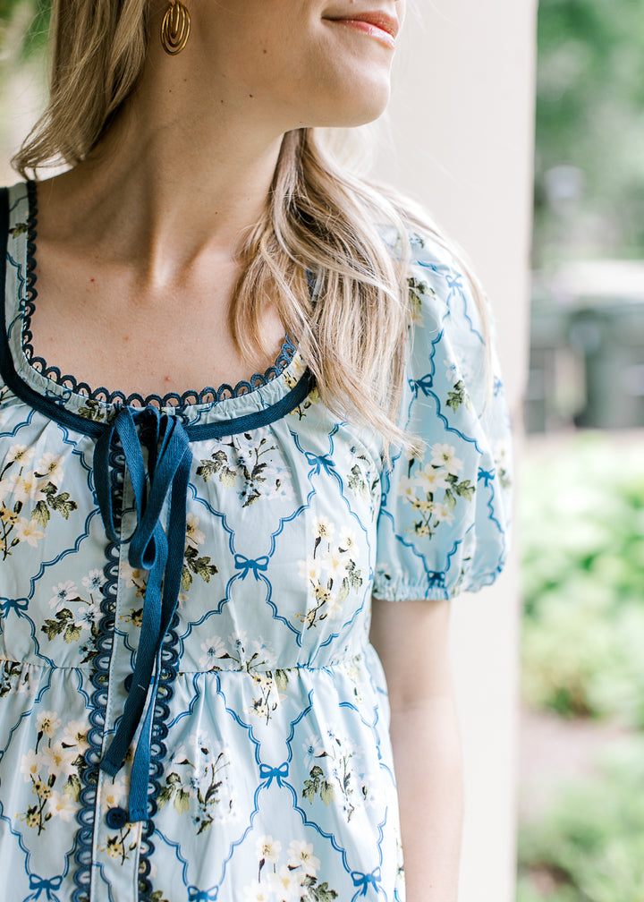 Close up of scalloped lace and faux tie on a baby blue top with cream floral and short sleeves.