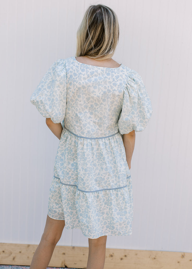 Back view of Model wearing a cream dress with blue flowers, v-neck and bubble short sleeves.