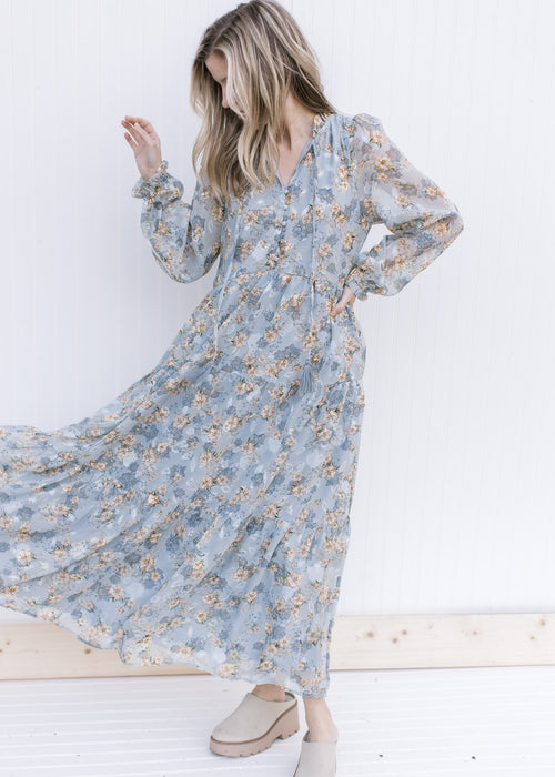 Model wearing a pale blue maxi with gold, cream and ivory floral, long sheer sleeves and  buttons.