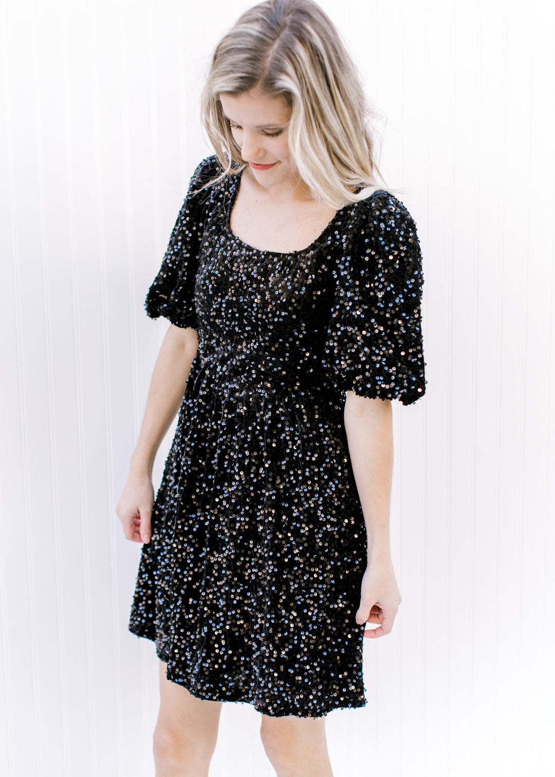 Model wearing an above the knee dress composed of black sequins, a square neck and 3/4 sleeves.
