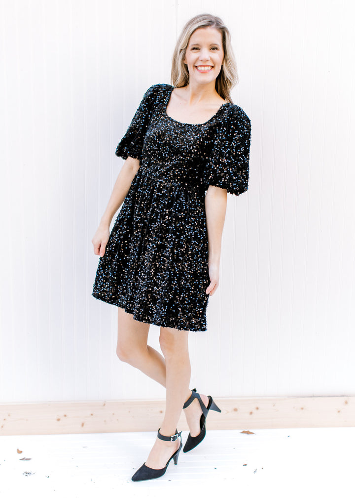 Model wearing heels with an above the knee dress with black sequins, square neck and 3/4 sleeves.