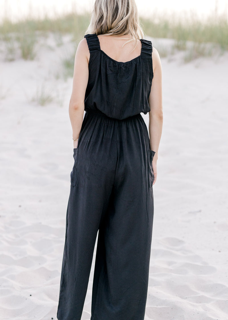 Back view of Model wearing a black wide leg jumpsuit with side patch pockets and elastic waist.