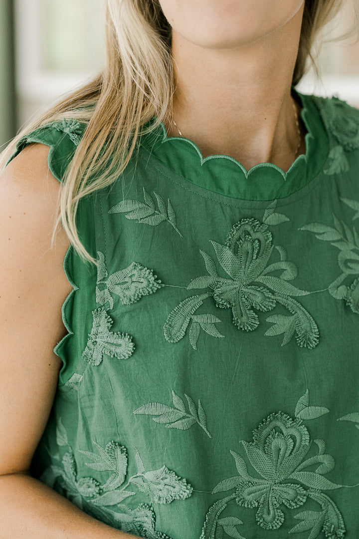 Close up of textured floral on a green, sleeveless dress with scallops at neck and arm. 