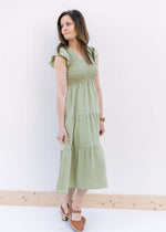 Model wearing heels with a basil green tiered midi with a smocked bodice and ruffle capped sleeves.