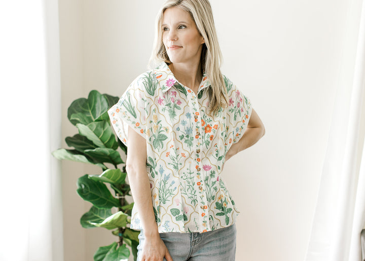 Model wearing a cream top with bright floral, button front, extended cap sleeves and pleated waist.