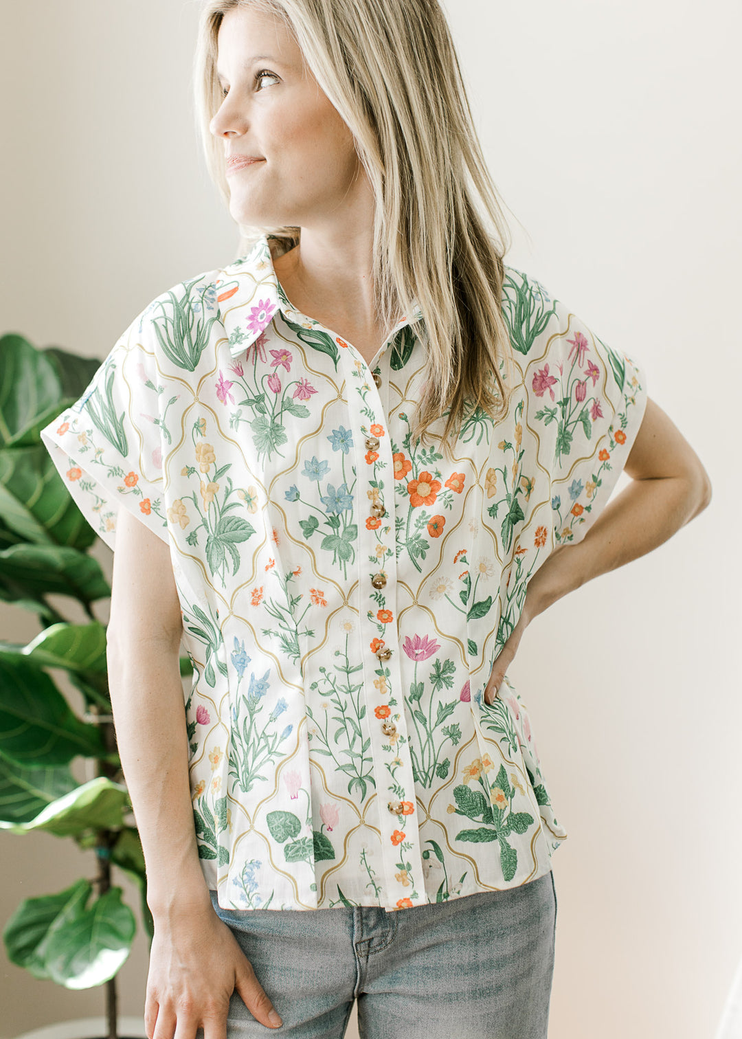 Model wearing a cream top with bright floral, button front, and pleating at the waist. 