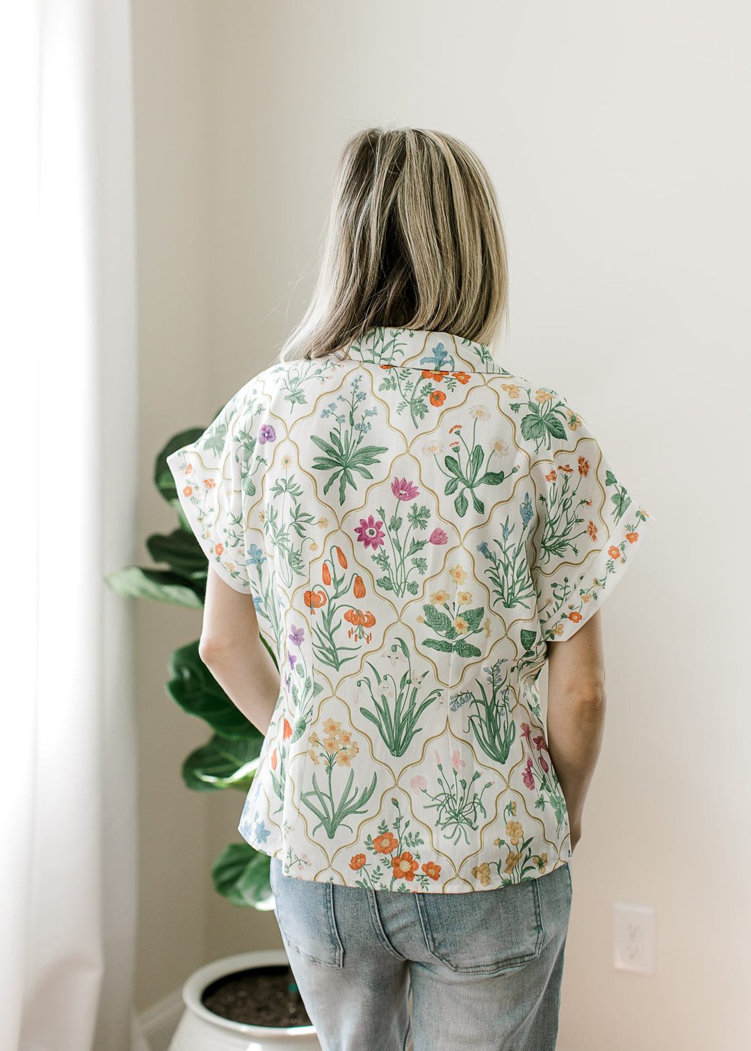 Back view of Model wearing a cream top with bright floral, extended cap sleeves and pleated waist.