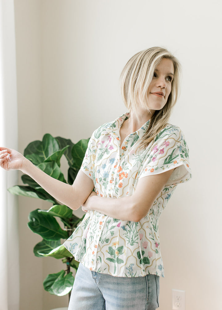 Model wearing a cream top with bright floral, button front, collar, and pleated waist.
