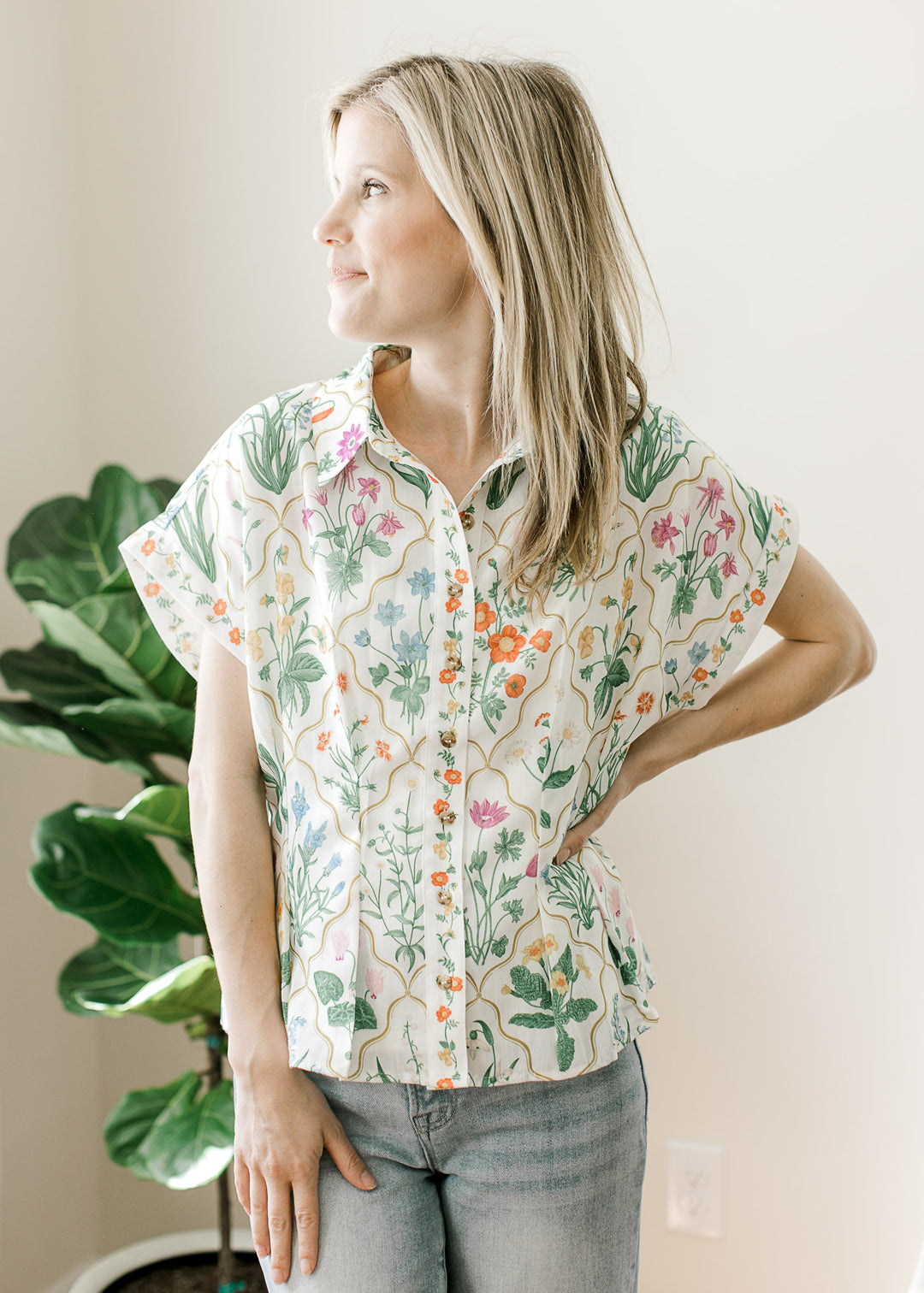 Model wearing jeans and a cream top with bright floral, button front, collar, and pleated waist.