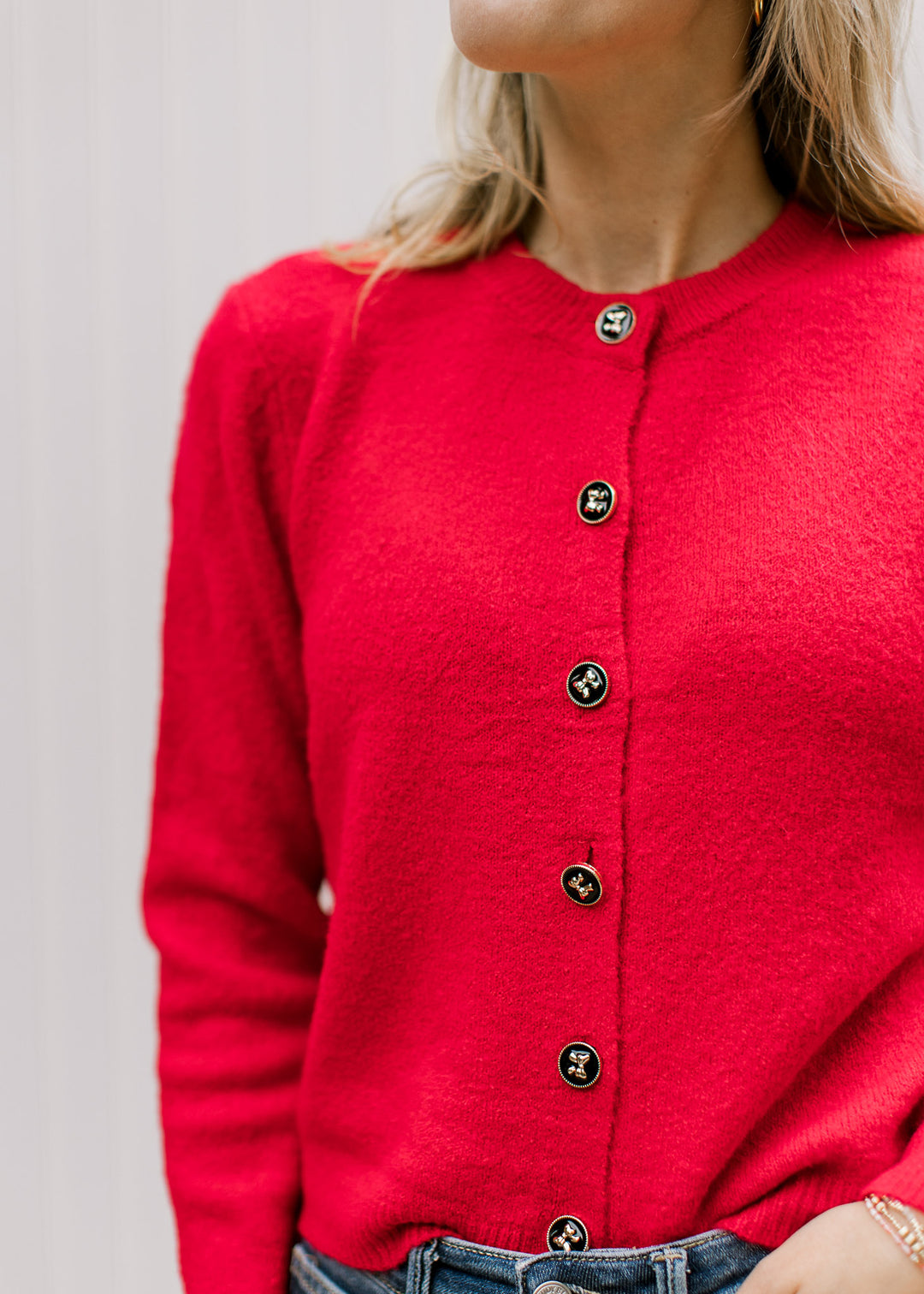 Model wearing a bright red cardigan with black buttons detailed in gold bows and a round neck. 