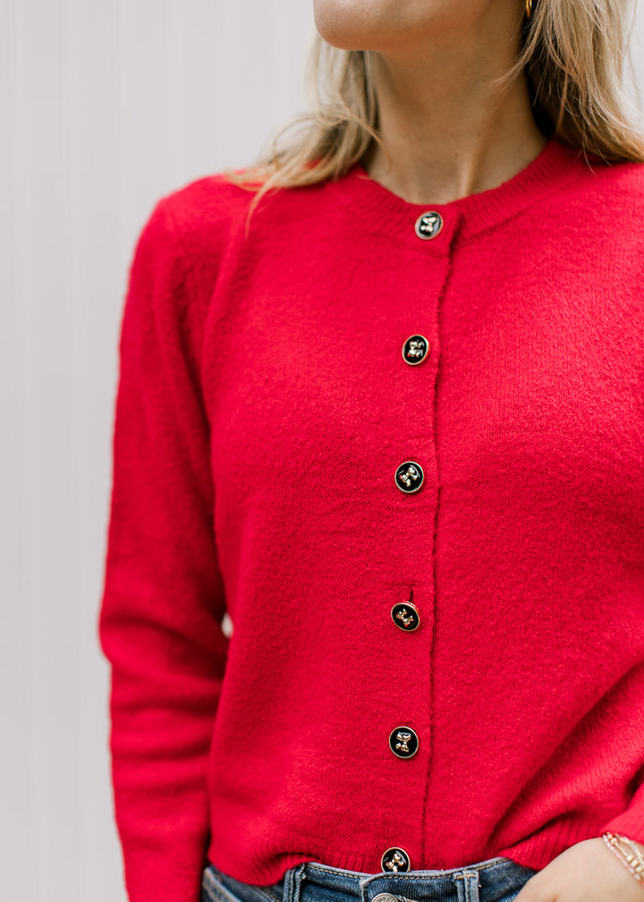 Model wearing a bright red cardigan with black buttons detailed in gold bows and a round neck. 