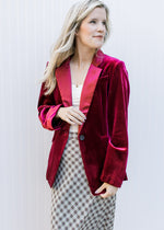 Model wearing a wine colored velvet blazer with long sleeves, faux pockets and flap collar. 