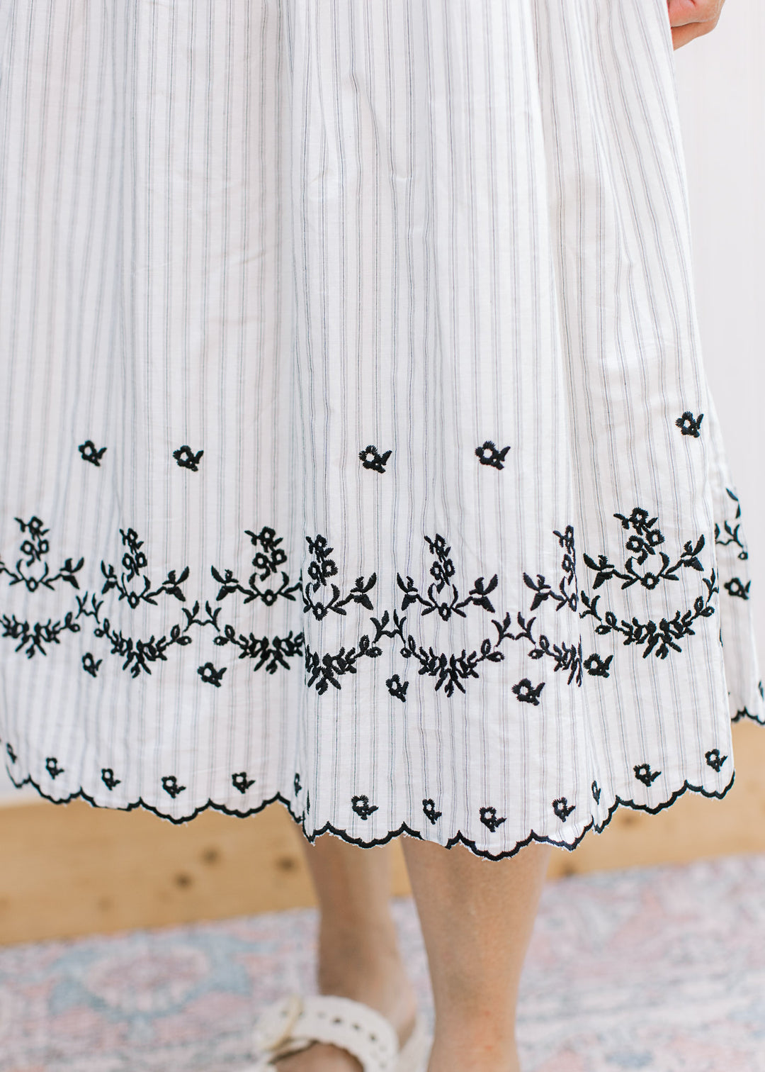 Close up of black floral embroidery and scalloped hem on a cream midi with gray stripes. 