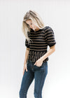 Model wearing a short sleeve black sweater with rust stripes and a scalloping at neck and sleeve. 
