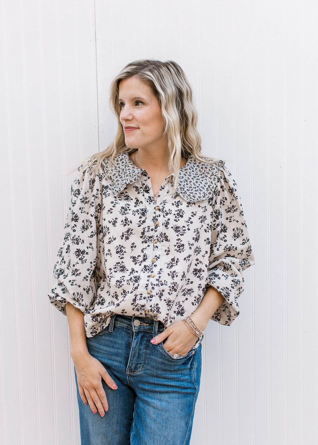 Model wearing a tan v-neck, button up top with black floral, long sleeves and exaggerated collar.