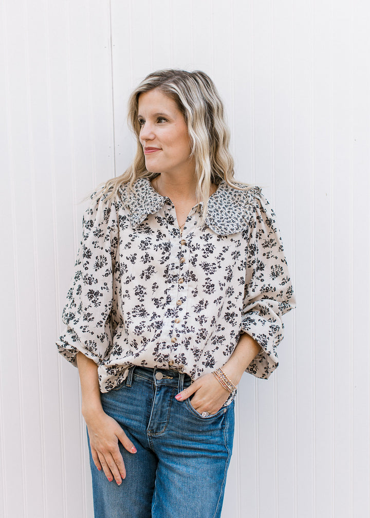 Model wearing a tan v-neck, button up top with black floral, long sleeves and exaggerated collar.