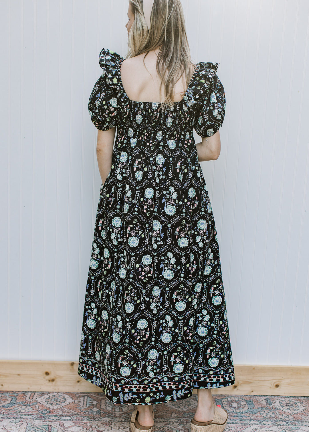 Back view of model wearing a short sleeve black maxi with floral, a smocked back and square neck.