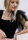 Close up of mesh ruffle sleeves on a black bodysuit with a square neck and double button closure. 