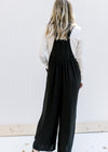 Back view of a Model wearing a black jumpsuit with two button adjustable straps and wide leg pants. 