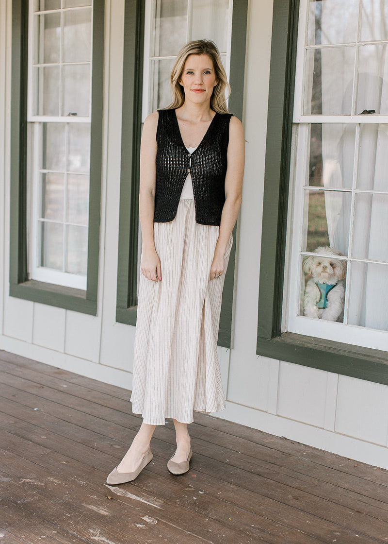 Model wearing a black vest over a sleeveless midi dress with a taupe skirt and black stripes.