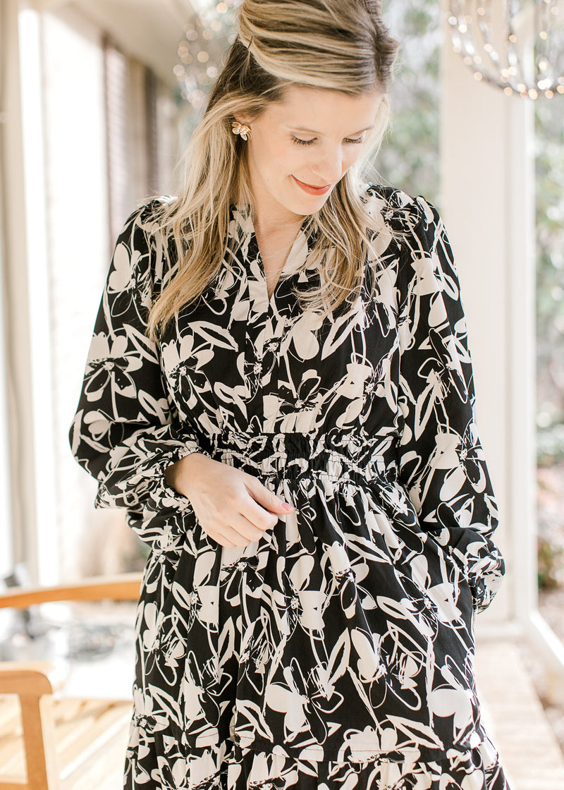 Model wearing a black dress with white flowers, smocked waist, pockets and long sleeves.