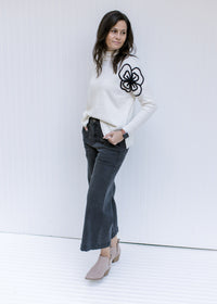 Model wearing a cream sweater with black embroidered flower detail, mock neckline and long sleeves.