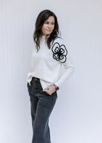 Model wearing a cream sweater with black embroidered flower detail, mock neckline and split sides.