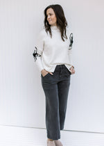 Model wearing jeans with a cream sweater with black embroidered flower detail and long sleeves. 