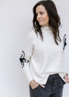 Model wearing a cream sweater with black embroidered flower detail, split sides and long sleeves.